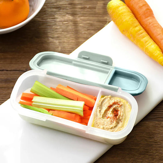 Bento box with compartments of carrots, celery, and hummus on a wooden table.