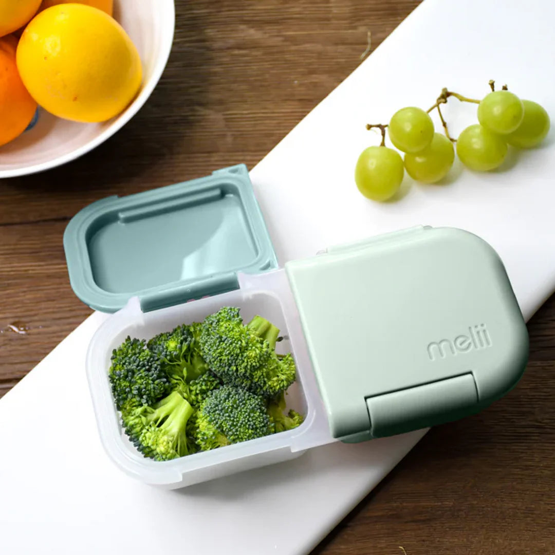 Green bento box with broccoli on a wooden table with fruits in the background