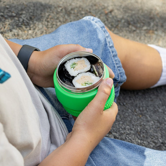 Person holding a green thermos with sushi inside, sitting on a concrete surface.
