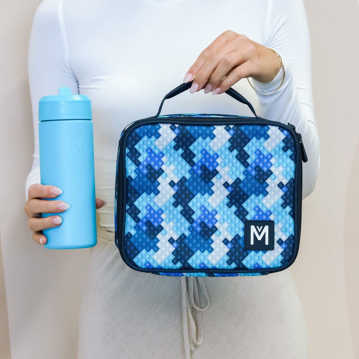 A person places a blue lunch container into an open blue patterned insulated lunch bag with a silver interior and a mesh pocket holding a name card, with a blue water bottle and a plant in the background.