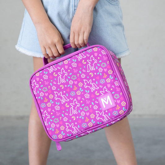 A person holds a pink unicorn magic lunchbag decorated with unicorns, flowers, hearts, stars, and moons, standing against a neutral background while wearing a light blue denim skirt.