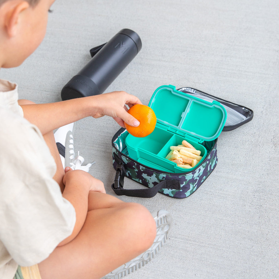 Child holding an orange next to a montiico mini stealth lunch bag opened to show snack containers inside for kids’ on‑the‑go use on a light gray surface