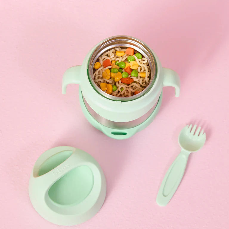 Mint green children's lunch container with food, lid, and fork on a pink background
