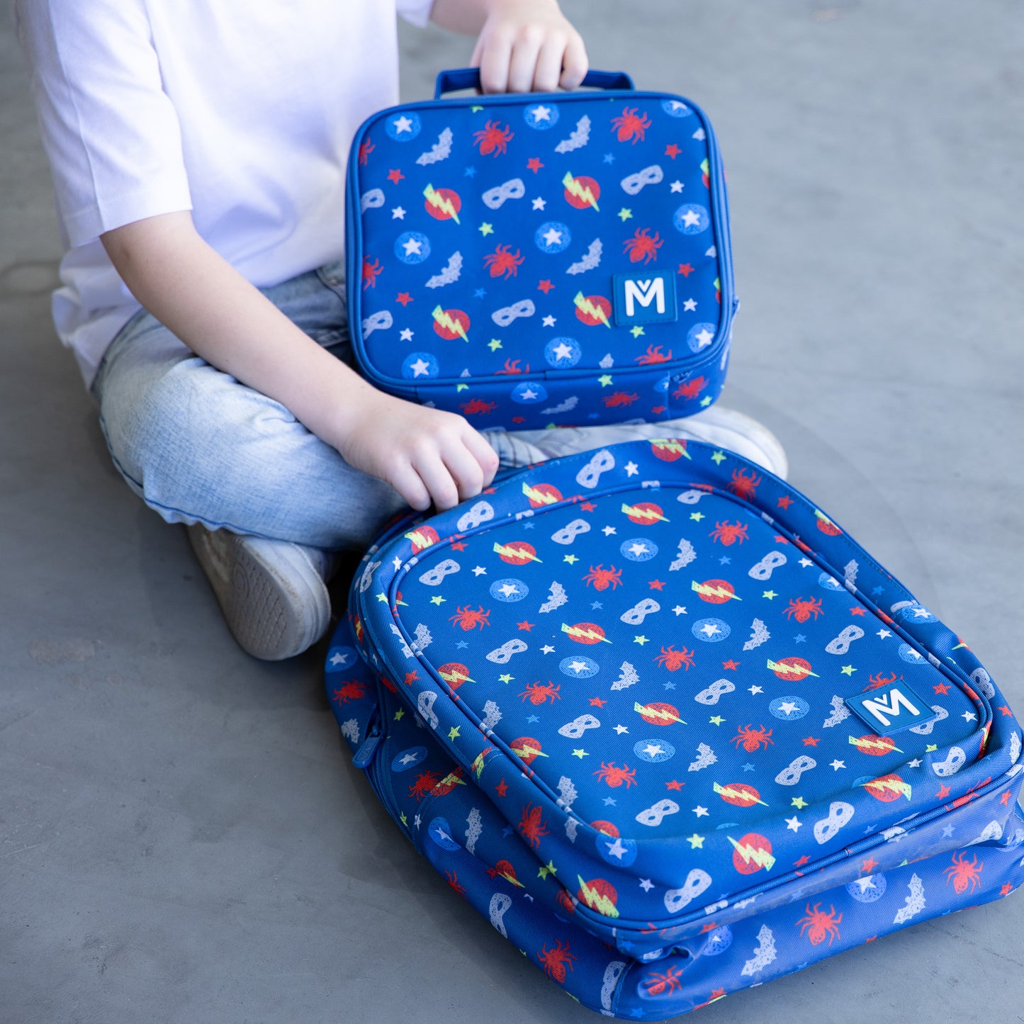 A young child wearing a white T‑shirt, light blue jeans, and white sneakers stands against a grey wall, holding a blue lunchbag with superheroes