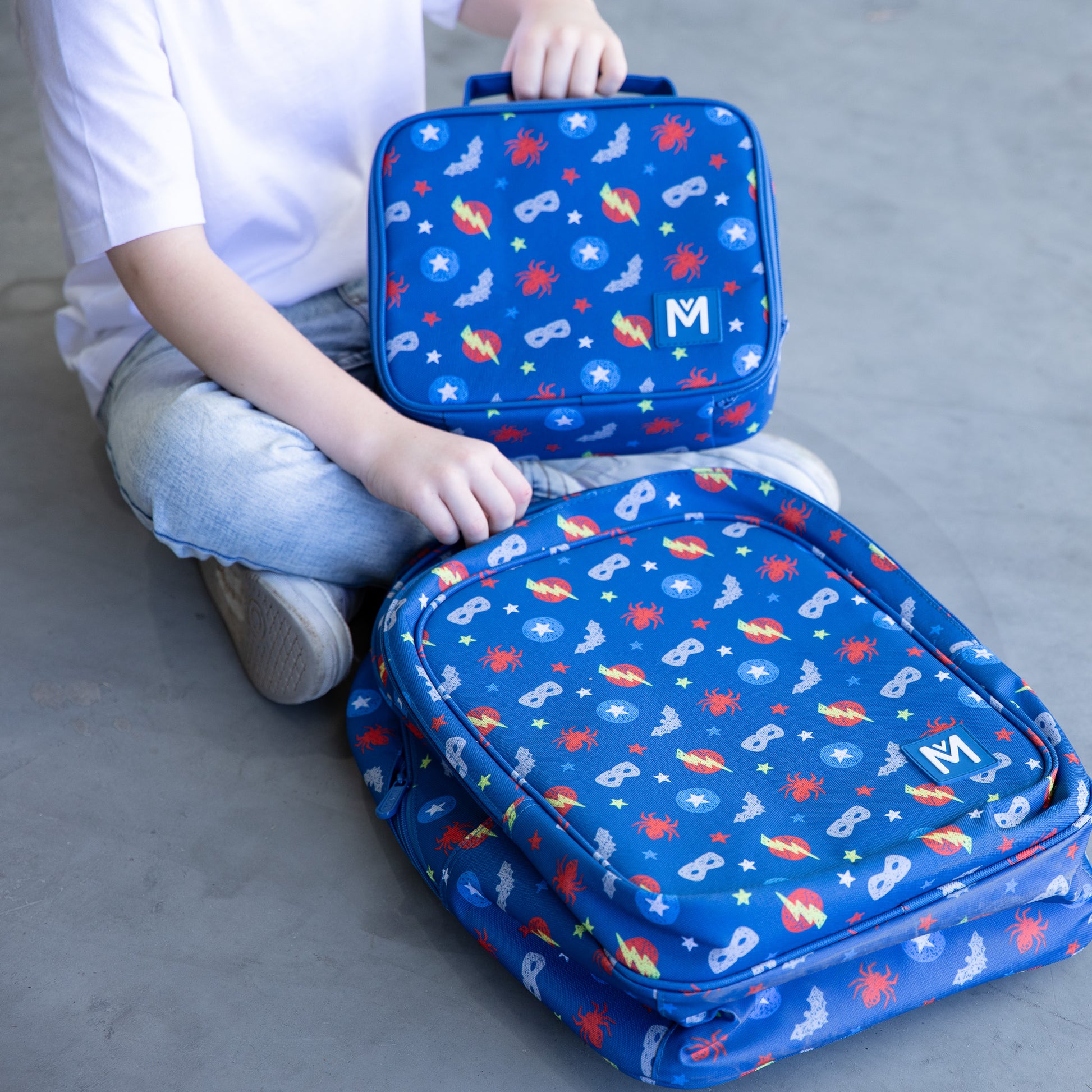 A young child wearing a white T‑shirt, light blue jeans, and white sneakers stands against a grey wall, holding a blue lunchbag with superheroes