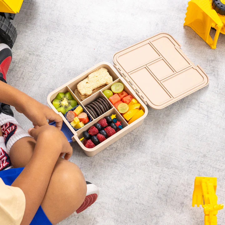Open MontiiCo Bento 5 lunch box packed with a sandwich and fresh fruit next to a toddler and toy construction vehicles.