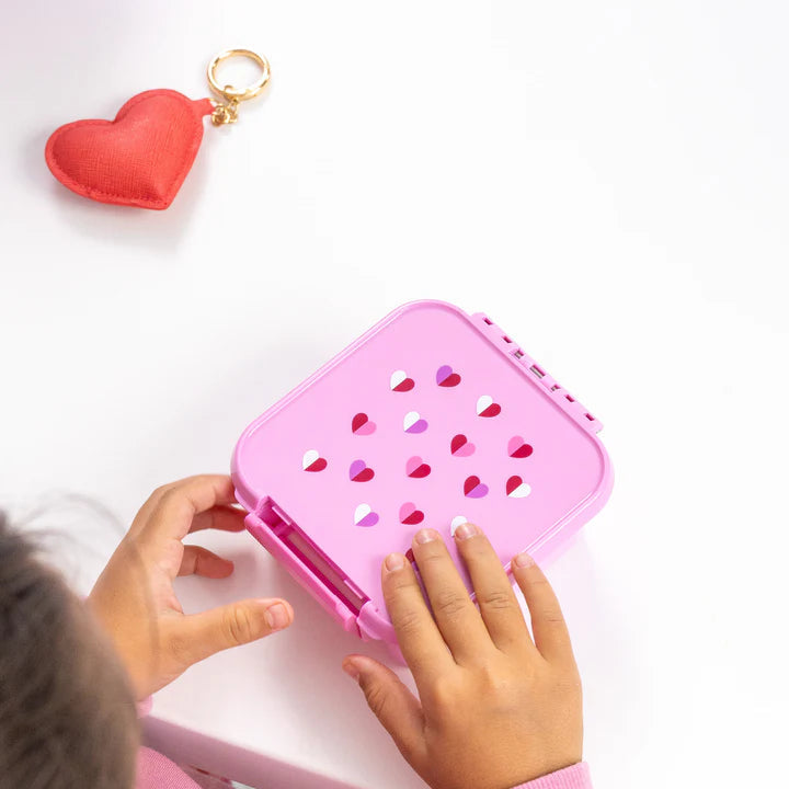 Child opening the pink MontiiCo Bento 2 Sweetheart snack box at a table next to a red heart keychain