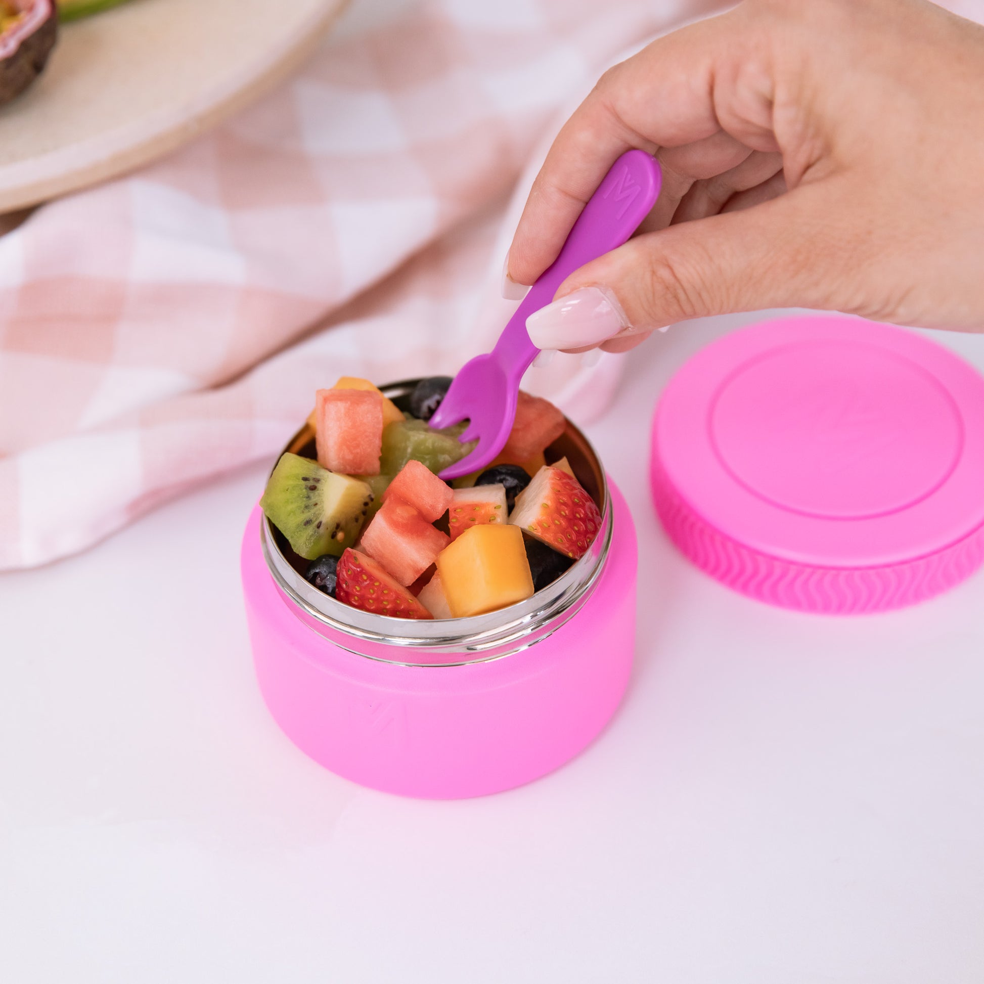 Pink snack container with fruit salad and a purple fork, on a light pink background.