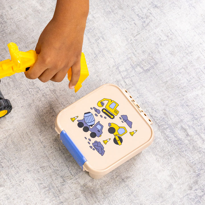 Overhead lifestyle shot of a child playing with toy trucks next to the closed MontiiCo Bento 2 Construction snack box.