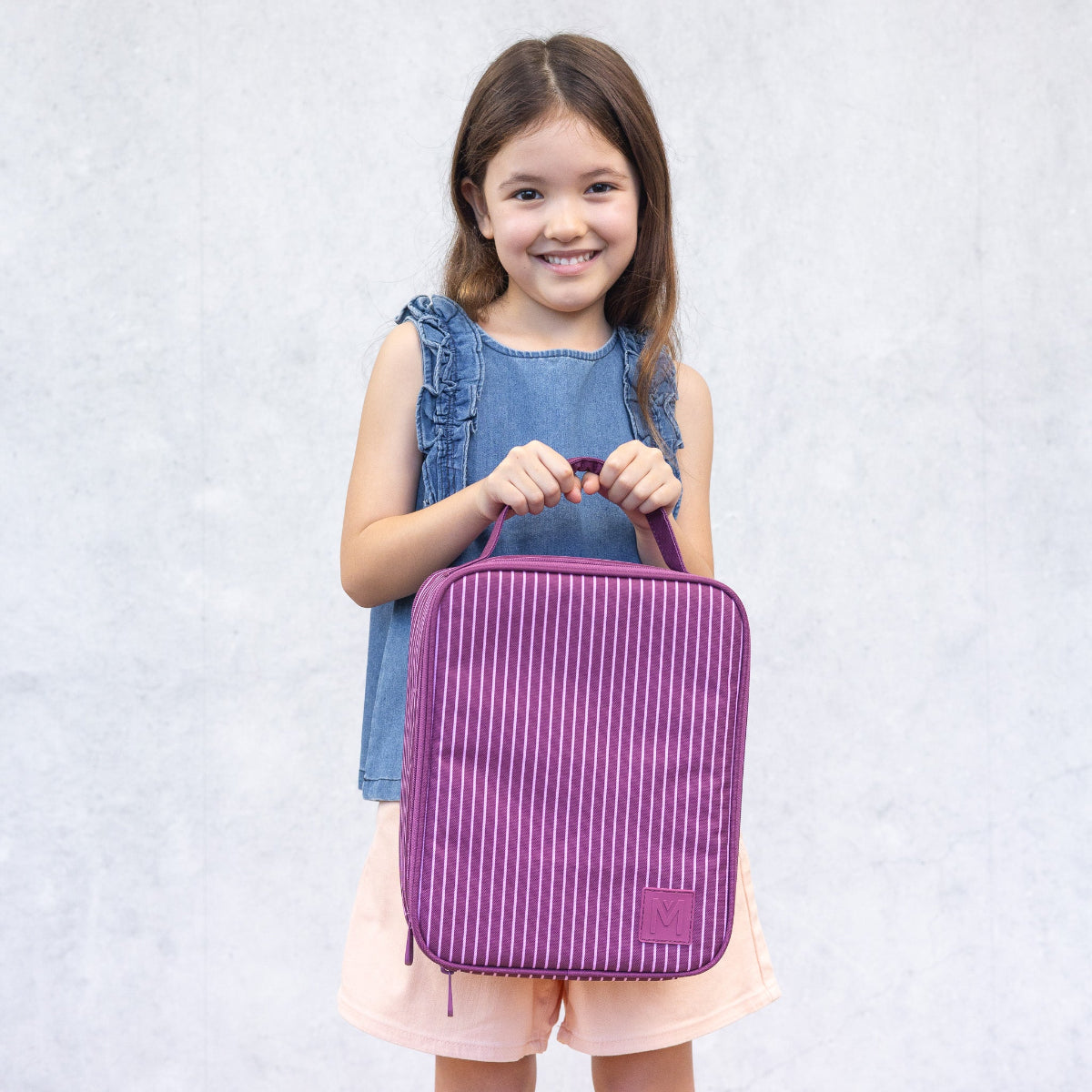 Young girl holding a purple striped Montiico large lunch bag against a light gray background