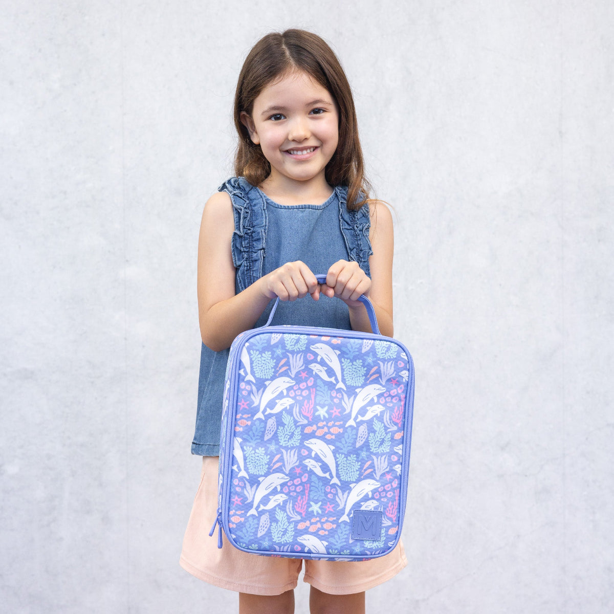 Young girl holding a blue lunch bag with dolphin design against a light gray background