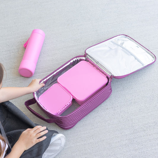 Montii Co Pink lunch box with compartments on a gray floor, child's hand reaching towards it.