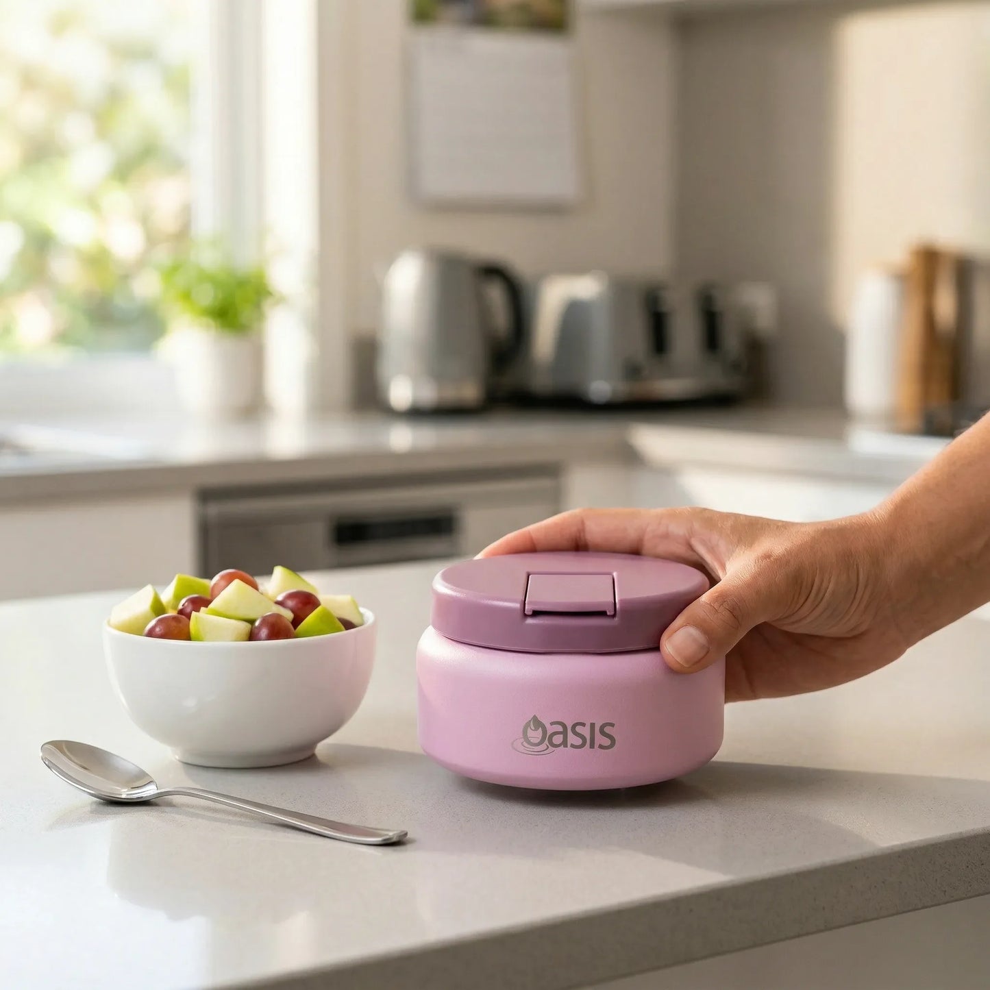 Person holding a pink 'Oasis' container on a kitchen counter with a bowl of fruit.