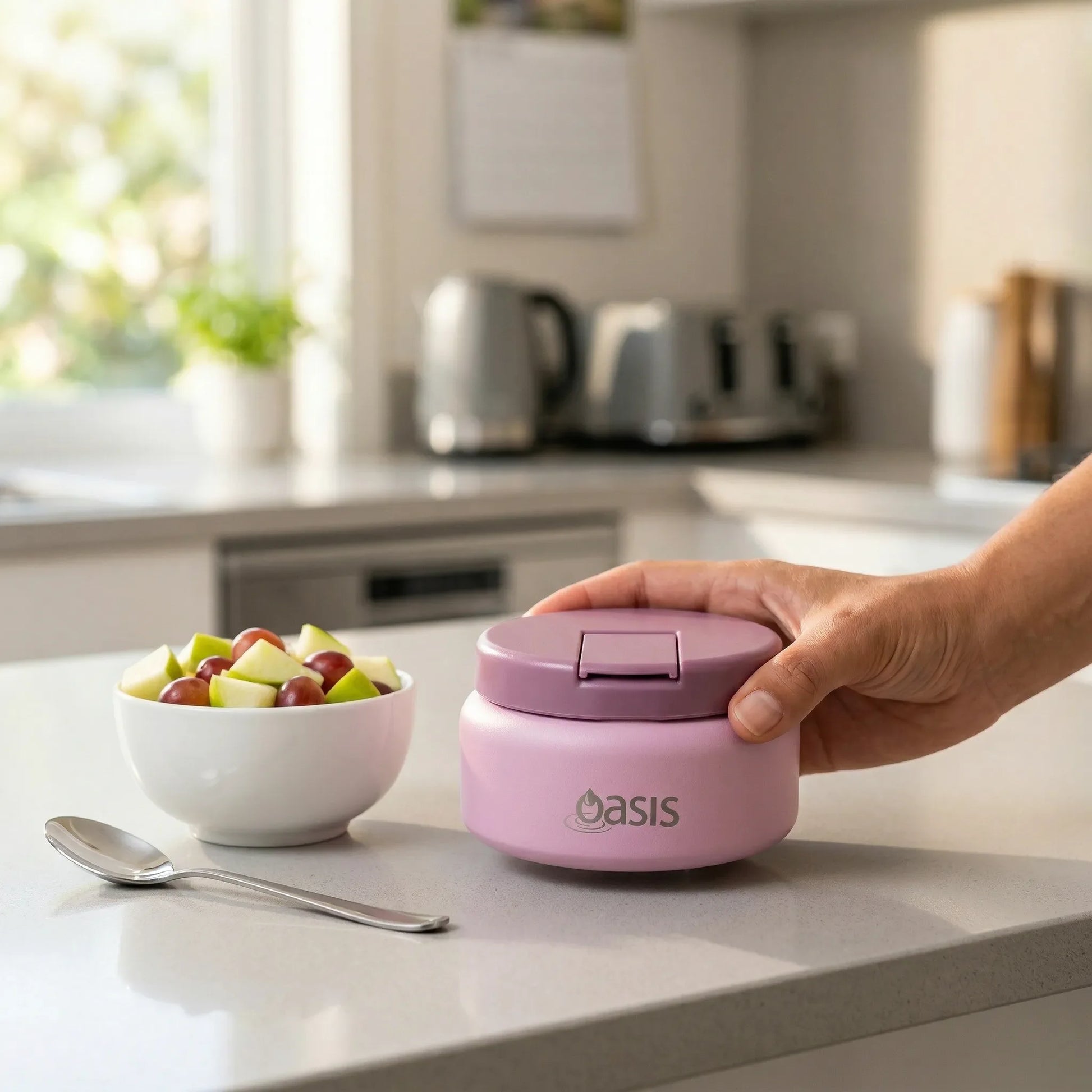 Person holding a pink 'Oasis' container on a kitchen counter with a bowl of fruit.