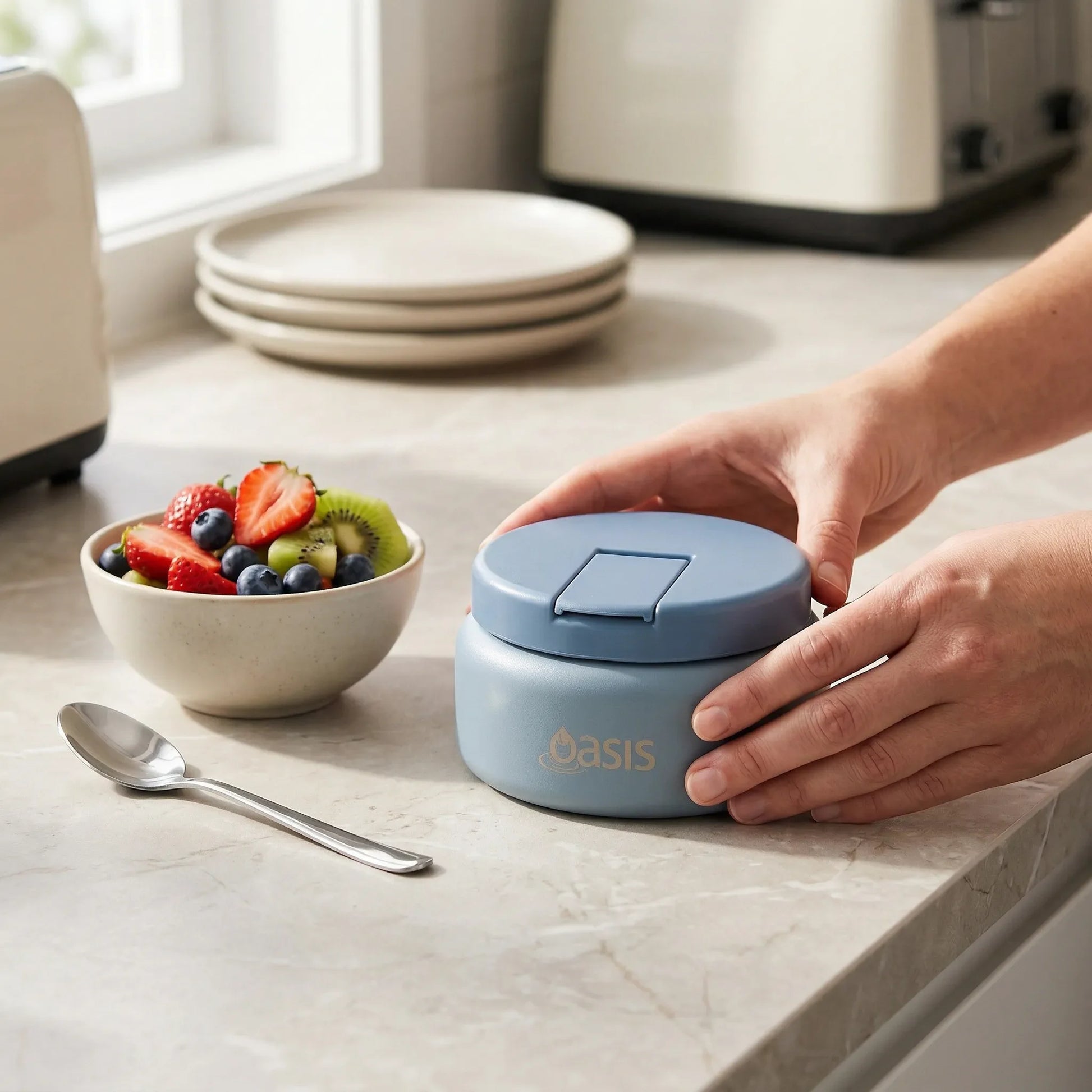 Person closing a blue 'Oasis' container on a kitchen counter with a bowl of fruit.