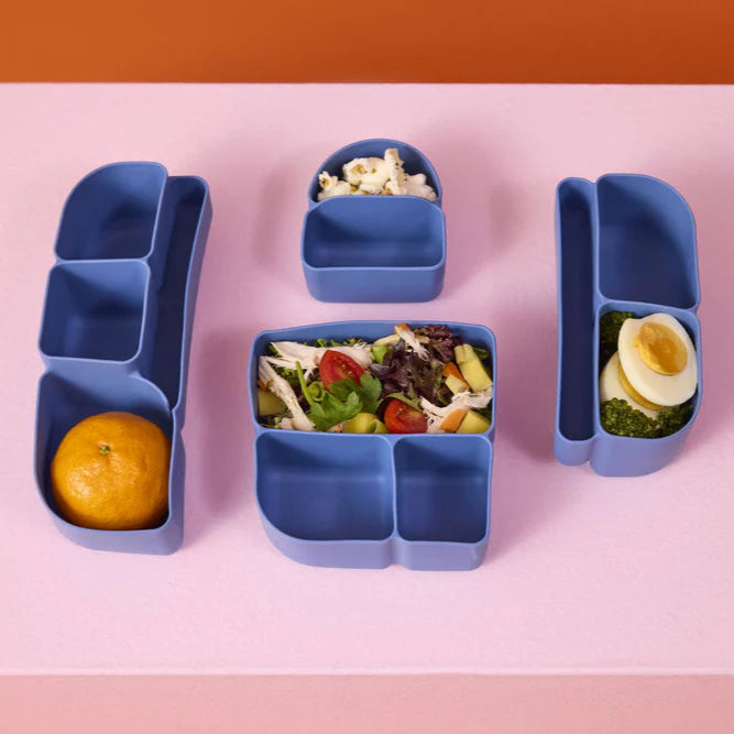 Blue bento box silicone inserts with compartments filled with food on a pink background