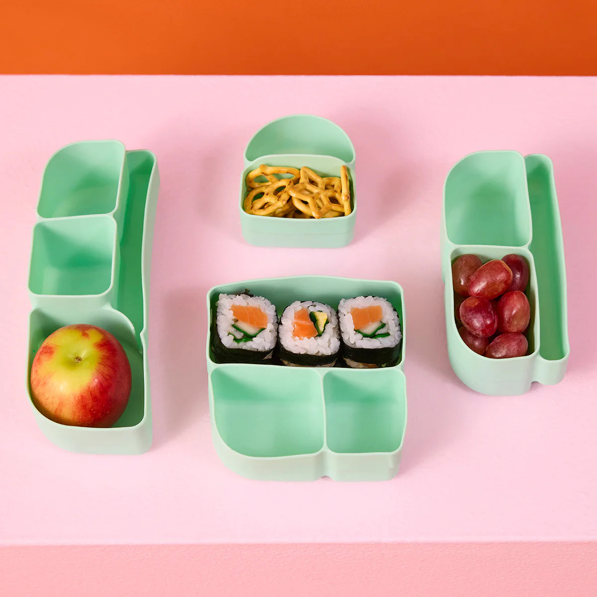 b.box Silicone Bento Tray : MINI - Forest Green bento box with compartments containing an apple, sushi, and grapes on a pink background