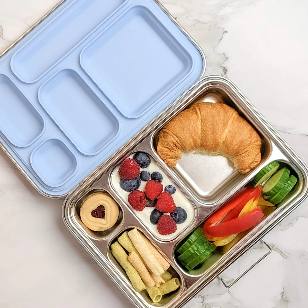 An open Ecococoon stainless steel leakproof bento box with five compartments, containing a variety of food items like fruits, vegetables, and a pastry. The box has a blue lid.