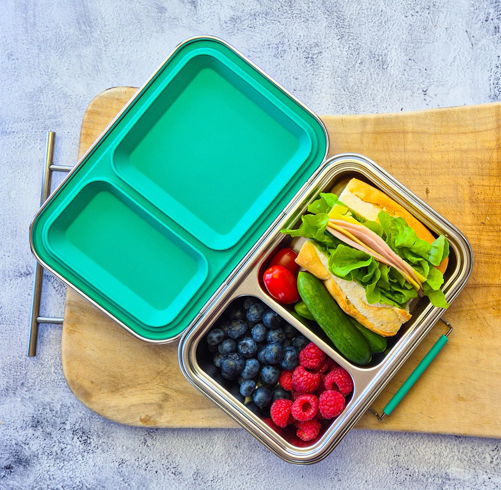 Ecococoon Metal lunch box with compartments containing a sandwich, fruits, and vegetables on a wooden board.