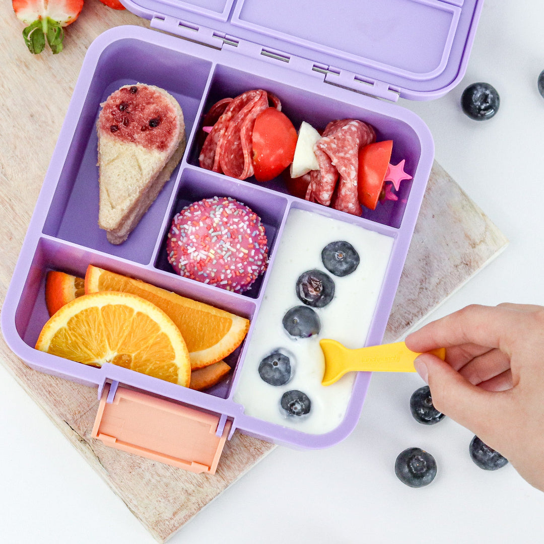 Child enjoying a Purple bento box meal  with Lunch-Punch-Fork-and-Spoon various food items on a white surface.