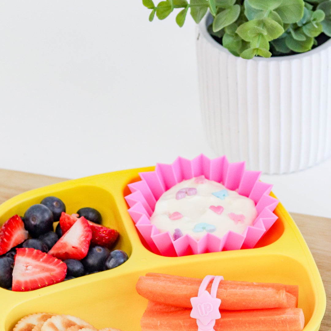 Blush jumbo silicone bento cups in Yellow divided plate with fruits, a dessert, and carrots on a white surface with a plant in the background.