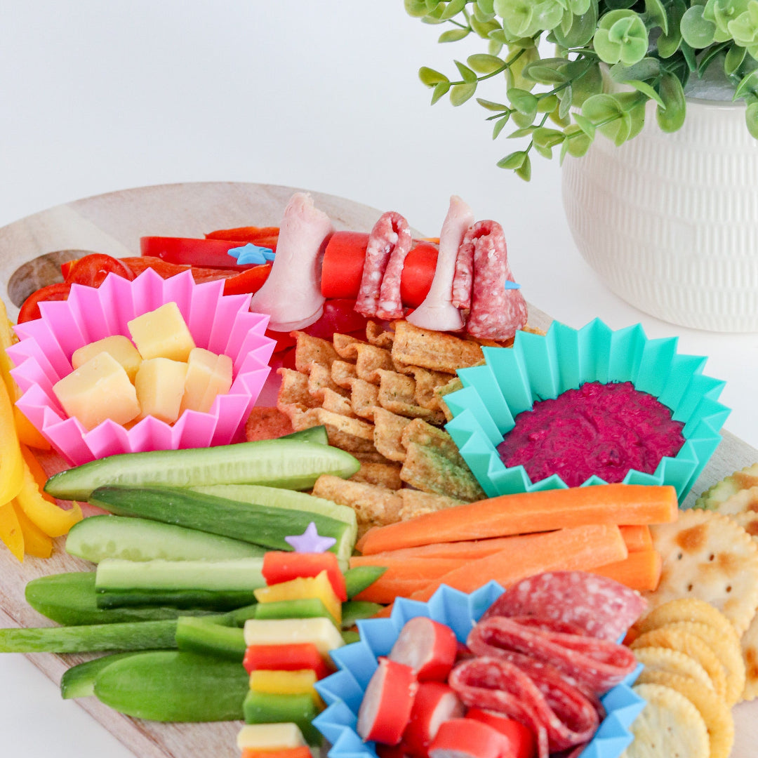 Jumbo silicone bento cups in burst colours, keeping snacks neat and fun inside assorted snacks including fruits, vegetables, and meats on a wooden board with a plant in the background.