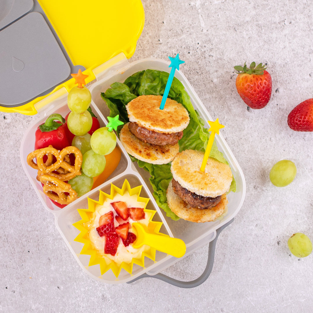 Children's lunchbox with mini burgers, fruits, and a sun-colored container decorated with lunch punch stix rainbow on a light gray background.