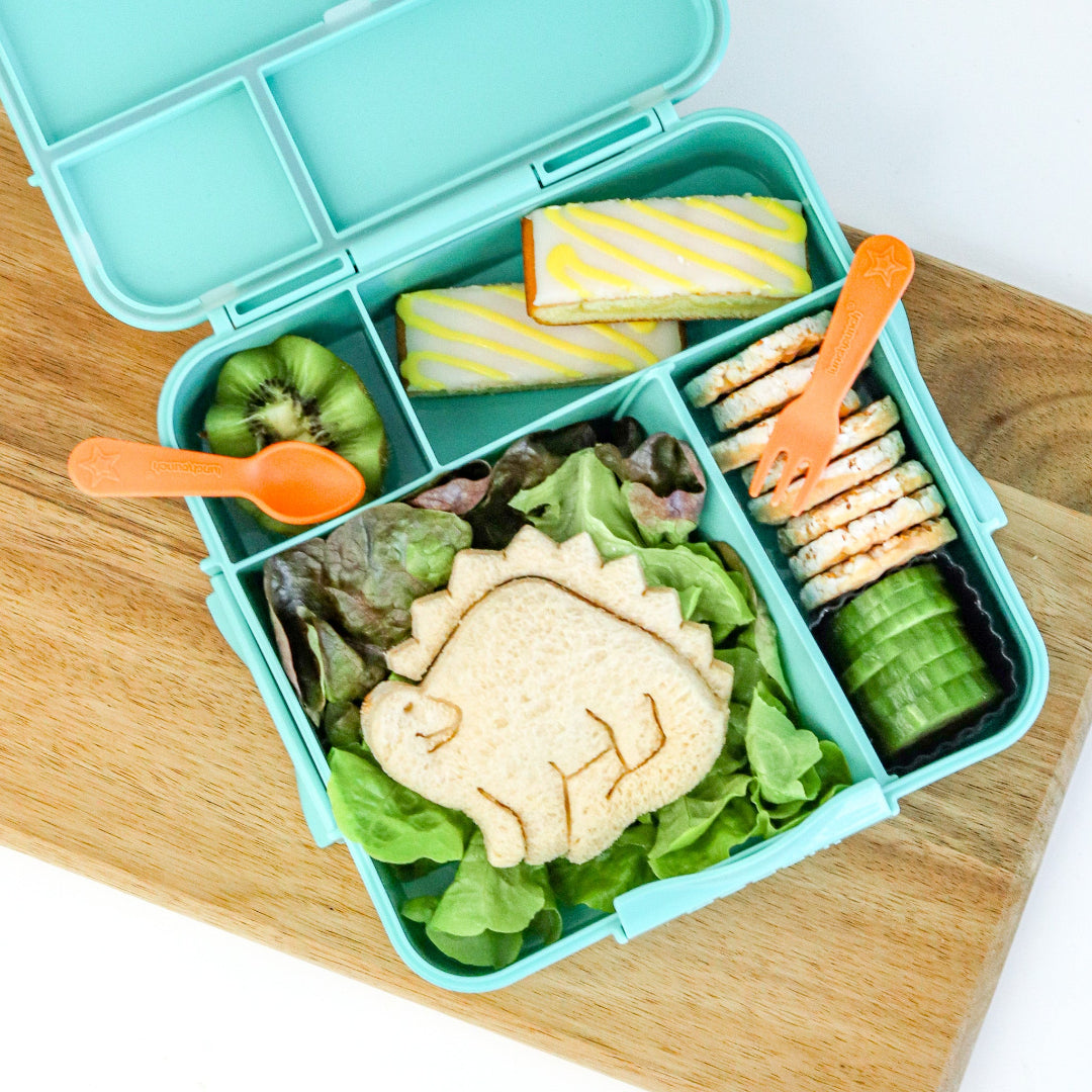 Children's lunchbox with dinosaur-themed sandwich, fruits, and snacks on a wooden surface, a lunchpunch cutlery set sits on the lunchbox