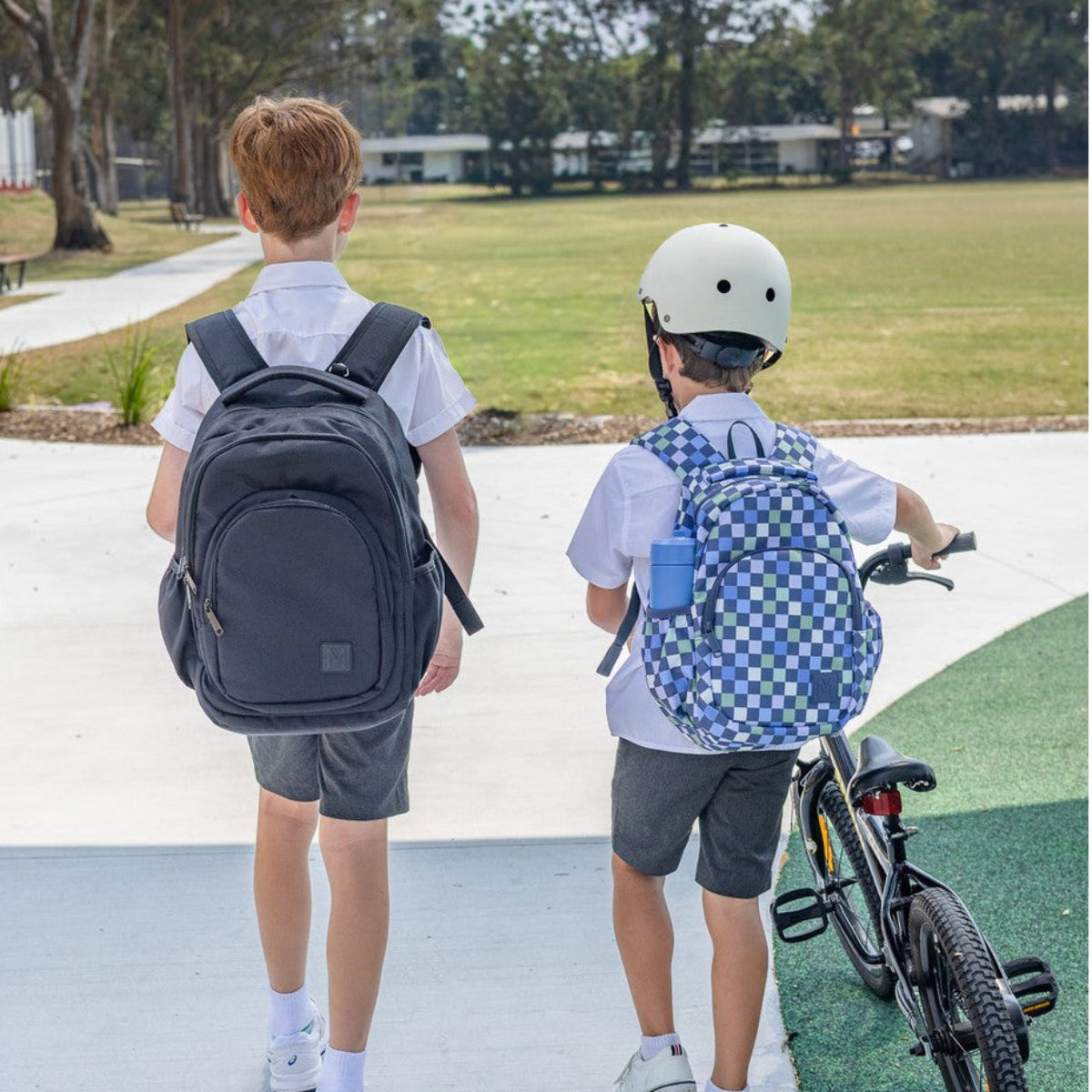 Two children, one with a backpack and the other with a bicycle, walking on a path outdoors.