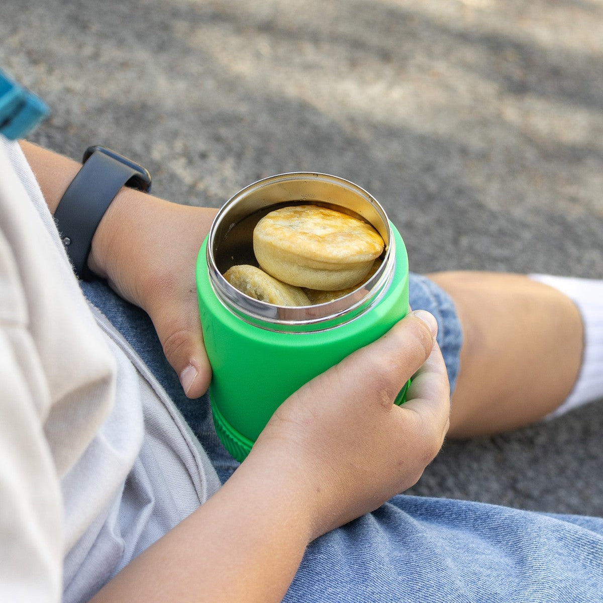Child holding a Montiico green thermos with cookies inside, sitting on a concrete surface.