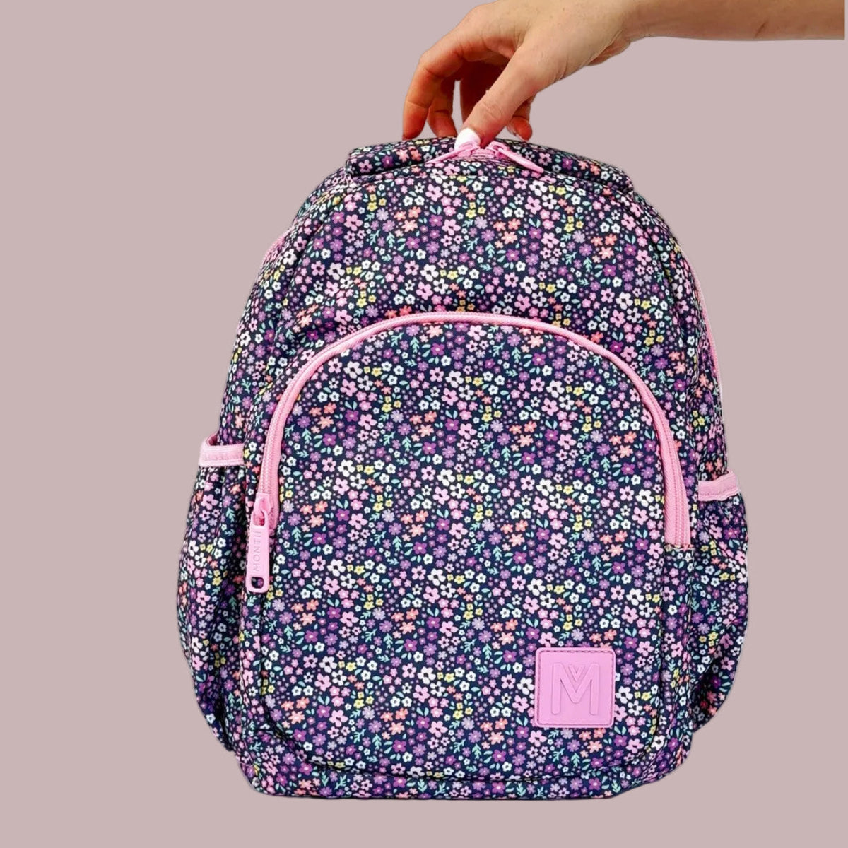MontiiCo Mini  Meadow Floral-patterned backpack with pink accents held by a hand on a white background