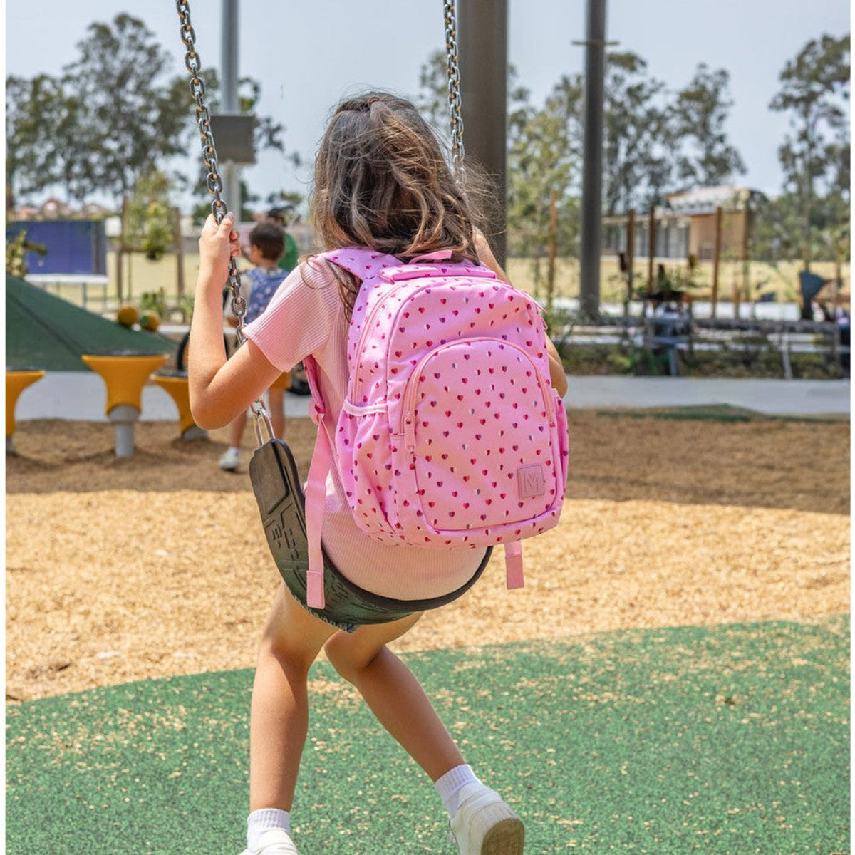 Child swinging on a playground with a pink sweet heart Monti backpack