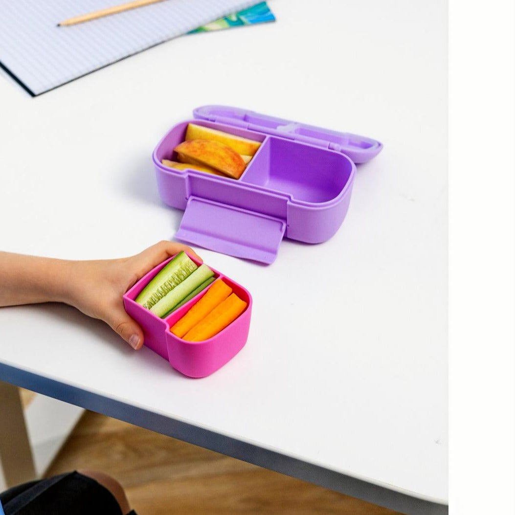 Purple MontiiCo Snack lunch box and pink Snack tray with snacks on a white table