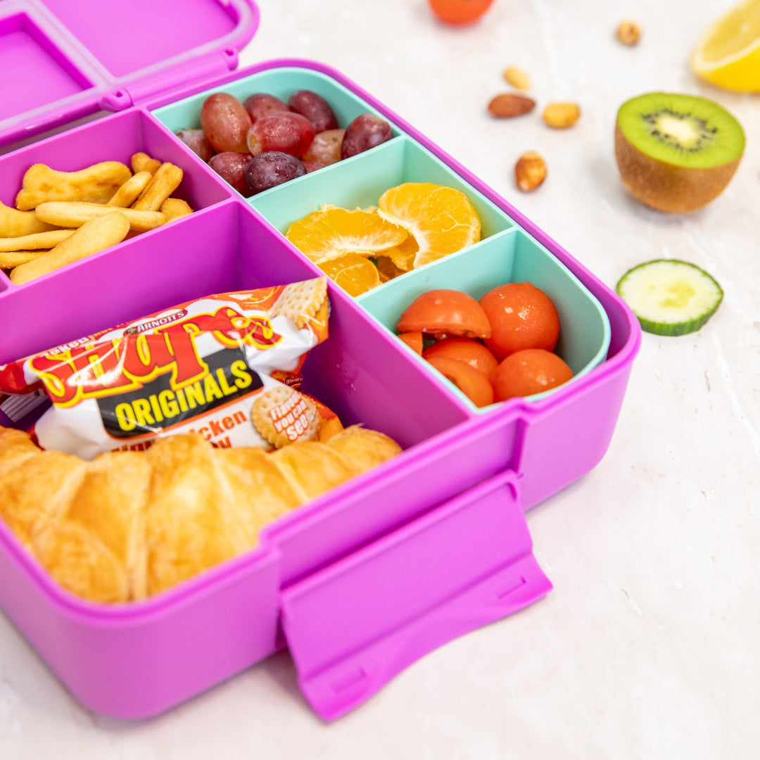 A fuchsia-colored Montii.Co Feast Bento Lunch Box with various foods arranged in its compartments, including fruits, vegetables, and a croissant.