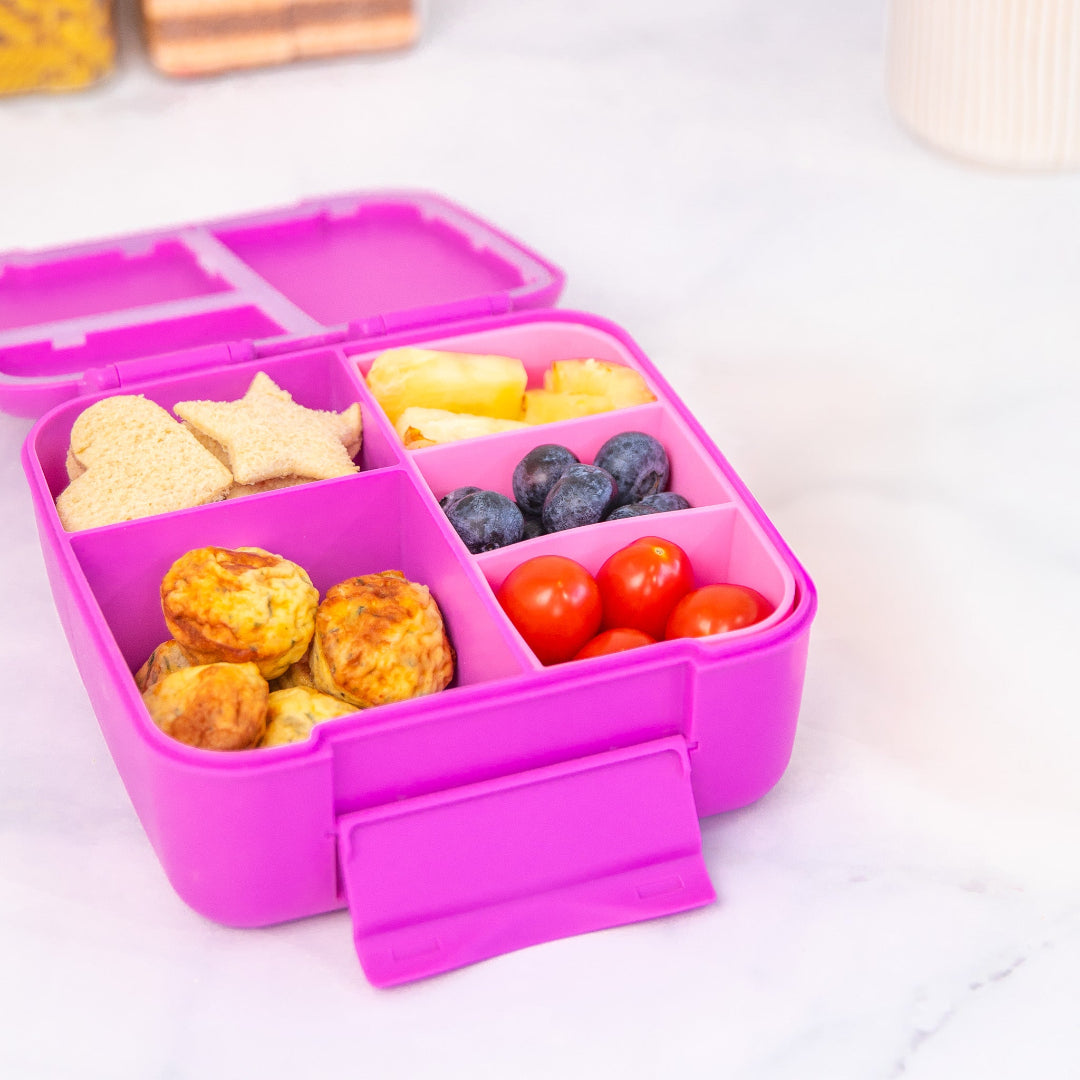 Kids’ snack tray by MontiiCo in Floss a silicone insert that fits inside a
Pink bento box with compartments filled with snacks on a white background