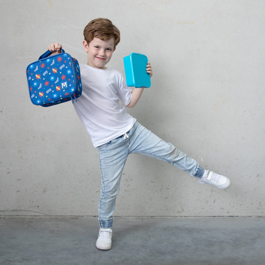 A young child wearing a white T‑shirt, light blue jeans, and white sneakers stands against a grey wall, holding a blue lunchbox with colourful cartoon designs in one hand and a turquoise book or tablet in the other, posing playfully with one leg lifted.