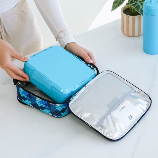 A person places a blue lunch container into an open blue patterned insulated lunch bag with a silver interior and a mesh pocket holding a name card, with a blue water bottle and a plant in the background.