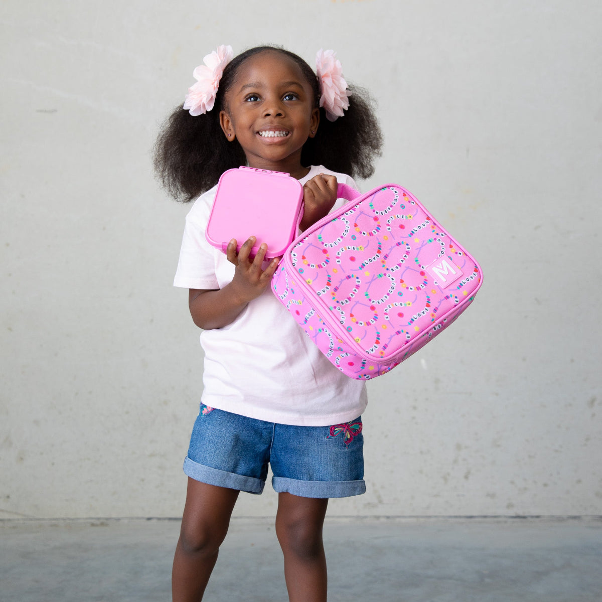 A young child wearing a white T‑shirt, denim shorts with embroidered details, and two large pink hair bows holds a pink lunchbox and matching container decorated with a colourful loop‑and‑dot pattern, standing against a plain background.
