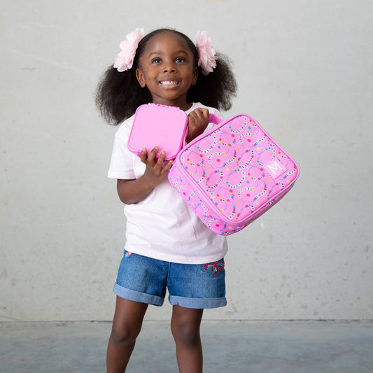 A young child wearing a white T‑shirt, denim shorts with embroidered details, and two large pink hair bows holds a pink lunchbox and matching container decorated with a colourful loop‑and‑dot pattern, standing against a plain background.