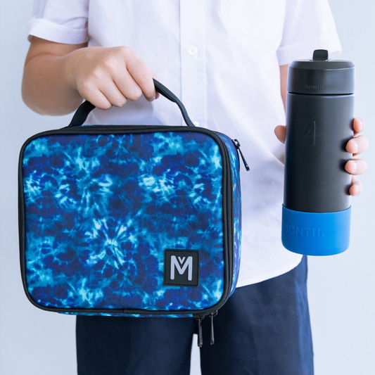 A person wearing a white button‑up shirt and dark pants holds a blue tie‑dye lunch bag with a black zipper and “M” logo, along with a black drink bottle featuring a blue base.