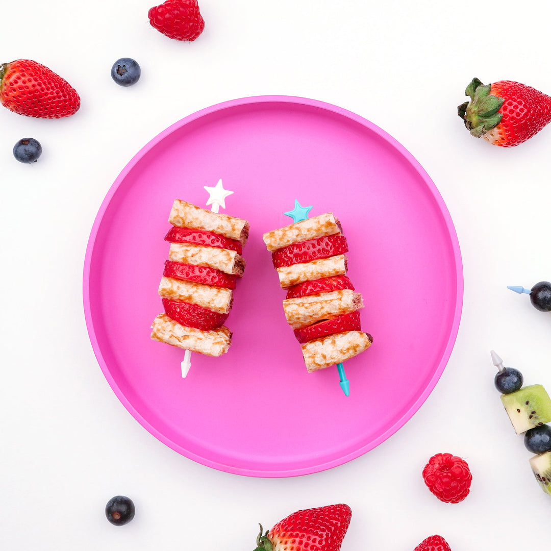 Two lunch punch stix skewers of fruit on a pink plate with strawberries, blueberries, and raspberries around.