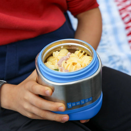 a person holding a yumbox stainless steel and blue insulated food jar labeled "Yumbox." The jar is filled with creamy rotini pasta, mixed with visible pieces of ham and green vegetables.
