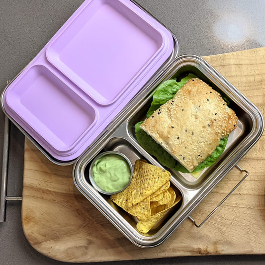 Ecococoon stainless steel leakproof bento box with two compartments and grape‑purple lid, the inner one purple, containing food items like a sandwich and snacks, placed on a wooden board.