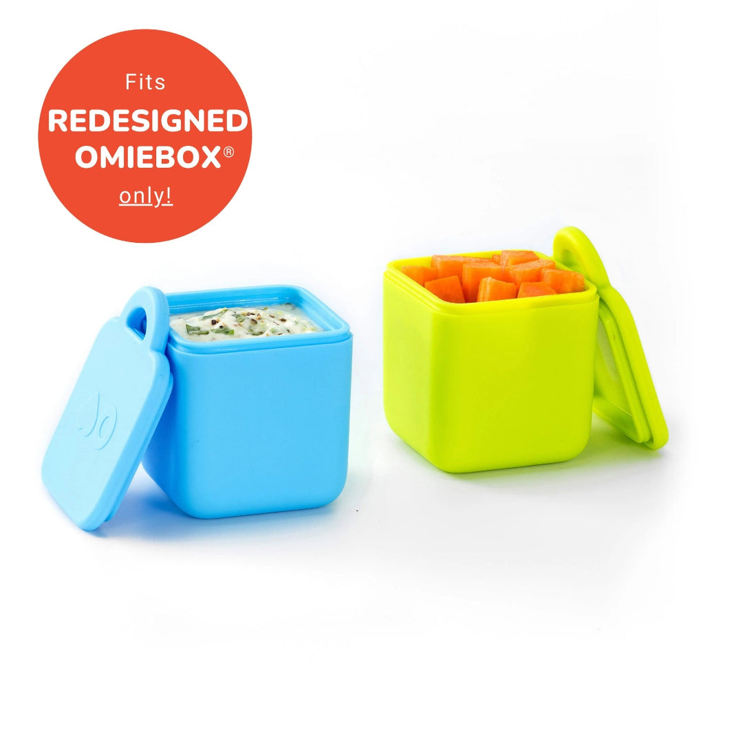 Two small, square food containers, one blue and one green, with lids, designed to fit inside the OmieBox V2. The blue container is filled with a beige substance, possibly a sauce or dip.