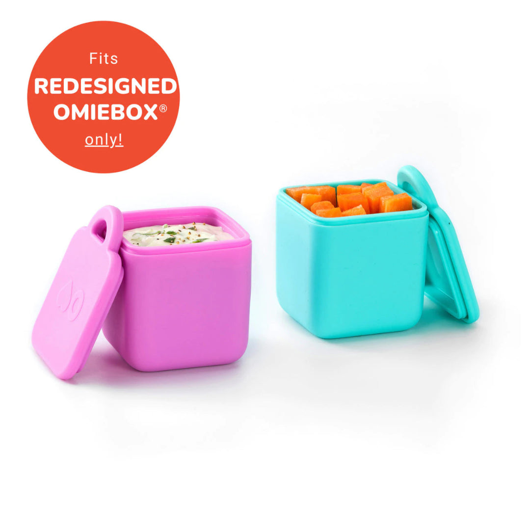 Two square silicone dip containers, one pink and one teal, designed to fit inside the OmieBox V2 lunchbox. The containers are shown open with dips inside, and the pink container has text indicating compatibility with the redesigned OmieBox V2 only.