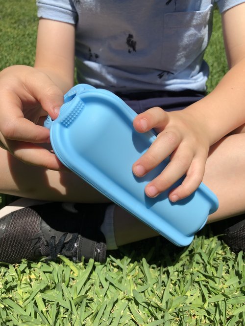 Child sitting outdoors holding a blue silicone reusable wrap holder, showing a flexible food holder designed to keep wraps and snacks secure and easy to eat.